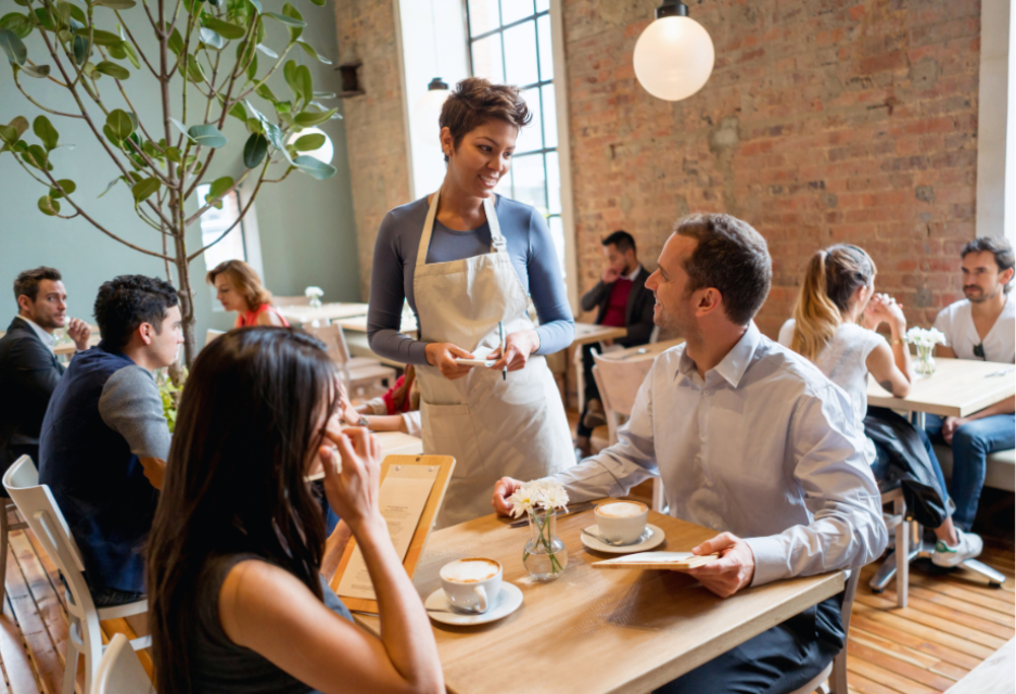 A busy, modern restaurant with a server assisting diners at a wooden table, surrounded by other guests, exposed brick walls, large windows, and warm contemporary lighting.